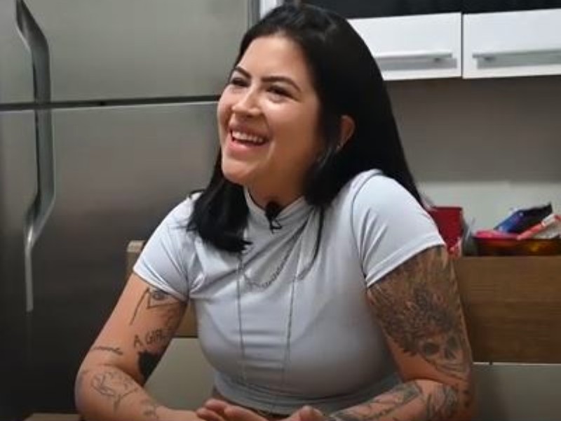 Nathalia está olhando de lado para a câmera, esboçando um sorriso. Ela tem os braços tatuados e usa uma blusa branca